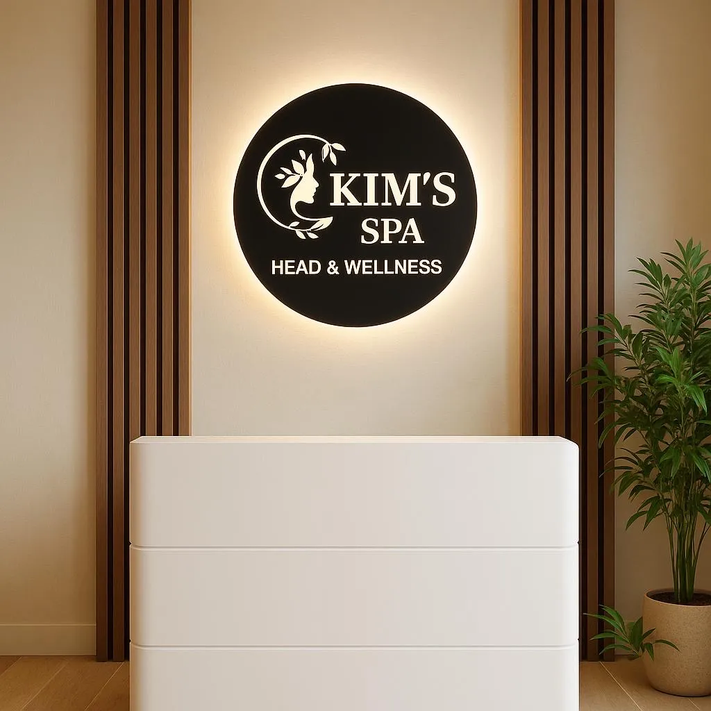 Moderner Empfangstresen in einem Spa mit beleuchtetem Logo 'KIM'S SPA HEAD & WELLNESS' an der Wand und grüner Pflanze daneben.