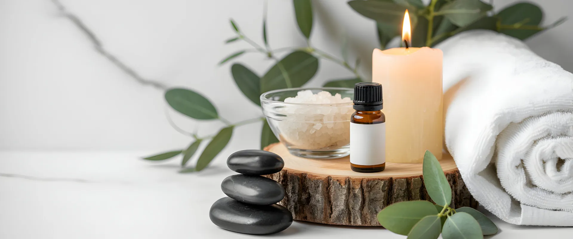 Hero-Header: Anordnung von Wellnessartikeln mit brennender Kerze, ätherischem Öl, Salz in Schale, weißen Handtüchern, schwarzen Massage-Steinen und grünen Blättern auf Holzunterlage.