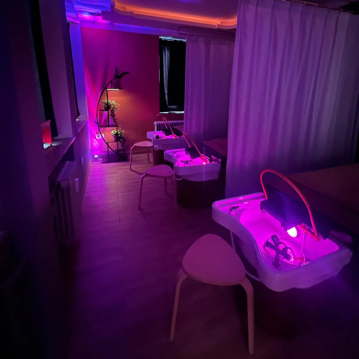 Beleuchteter Spa- und Salonbereich mit rosa-violettem Licht, weißen Waschtischen, roten Vorhängen und Pflanzenregal.
