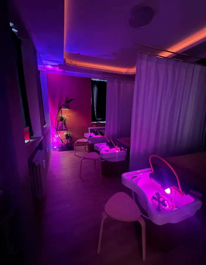 Beleuchteter Spa- und Salonbereich mit rosa-violettem Licht, weißen Waschtischen, roten Vorhängen und Pflanzenregal.
